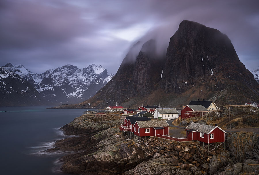 A delicate blanket of clouds caresses mount Festhæltinden, rising above the Atlantic waters. The red houses are traditional fishermen's cabins, now renovated and converted into accommodations.